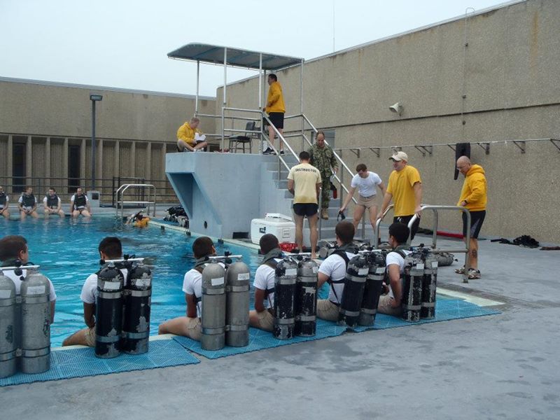 A group of men sitting on the side of a swimming pool with oxygen tanks receiving instructions from someone