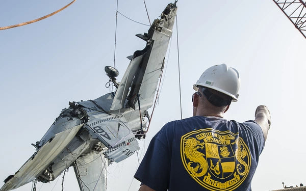 Man in a hard hat directing a crane that's lifting a crashed military jet.