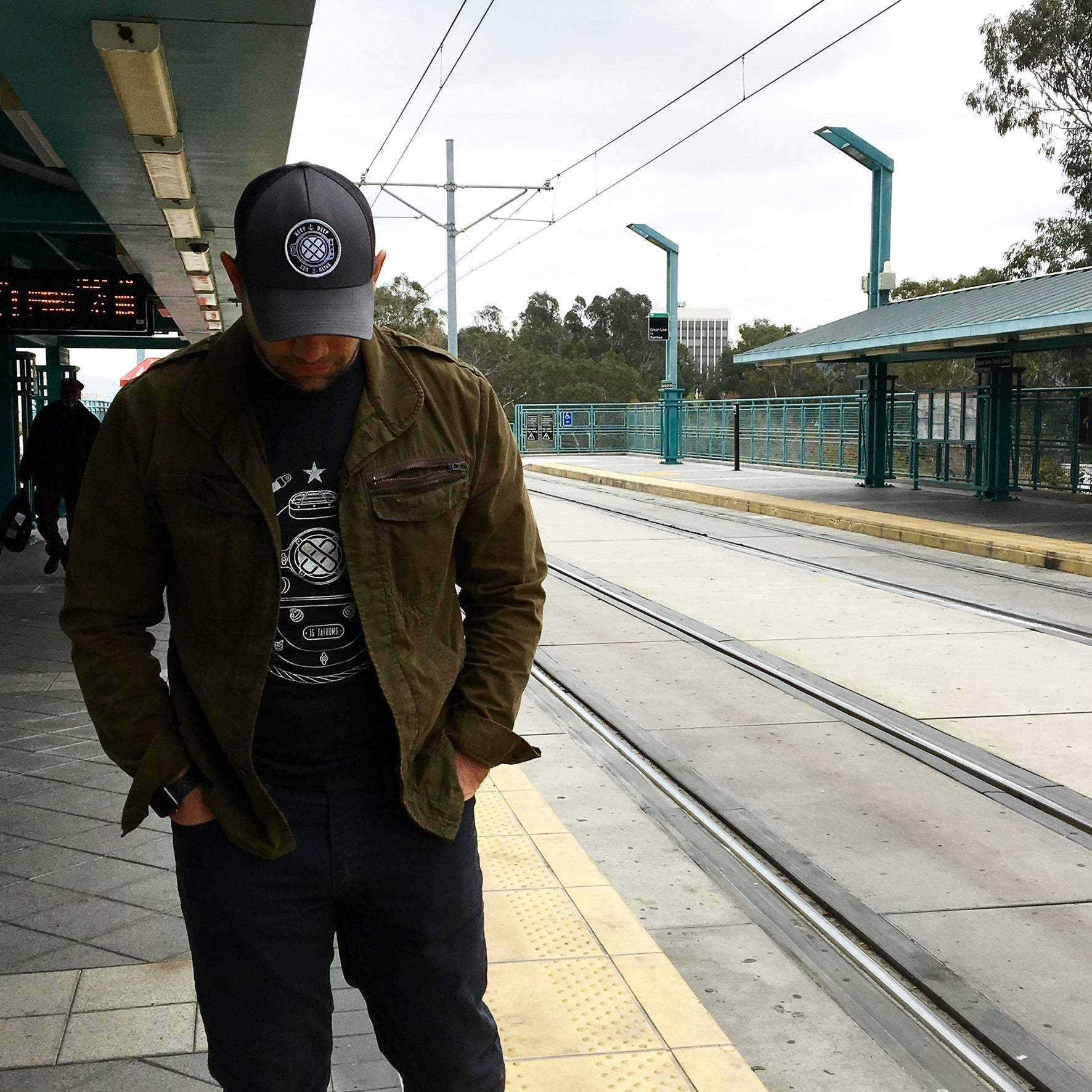 Man standing at a train station wearing black 15 Fathoms hat and tee