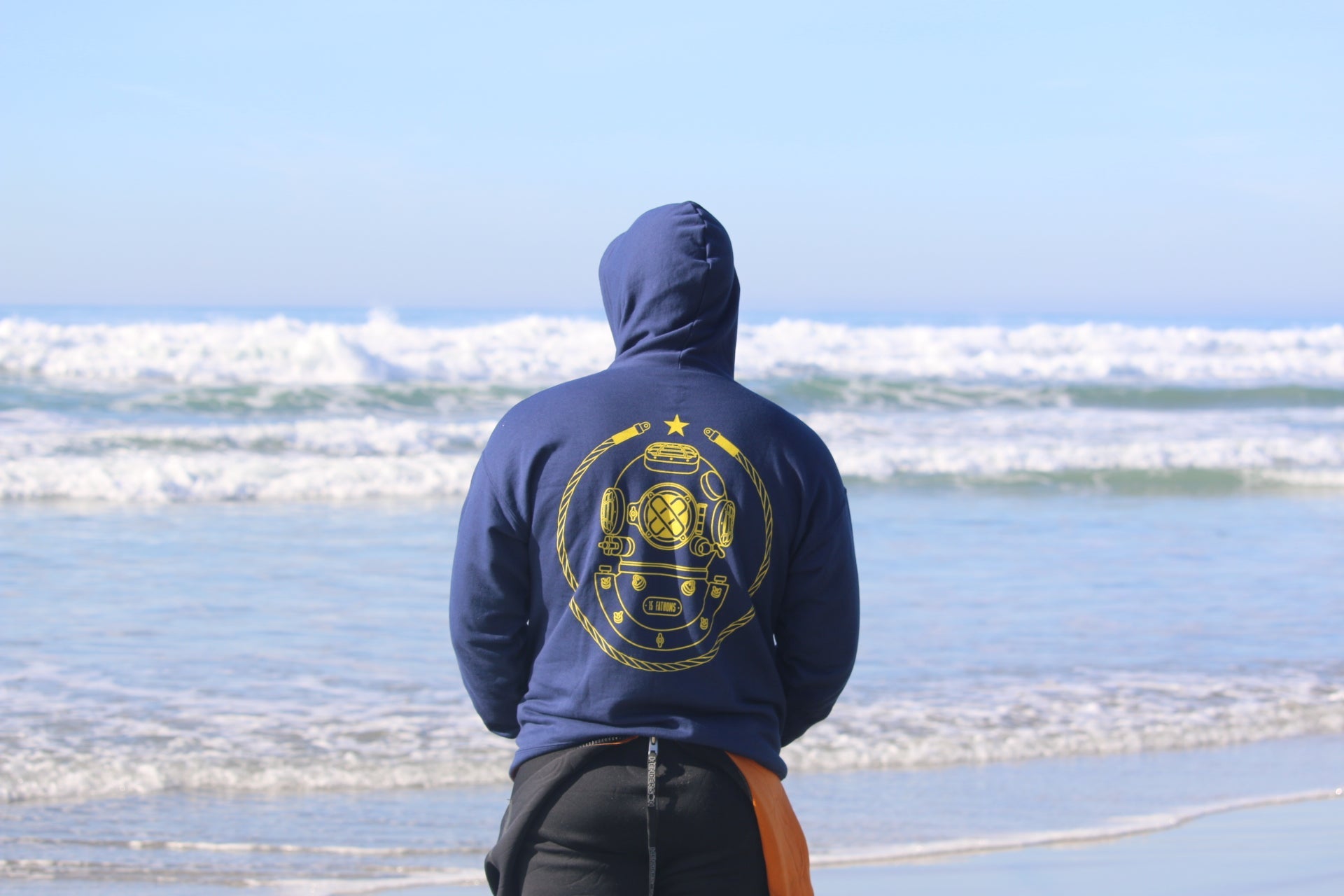 Man standing on the beach looking out into the ocean wearing navy blue 15 Fathoms hoodie