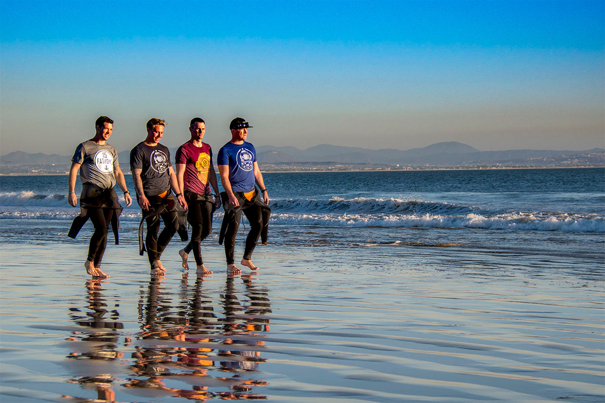 Four men walking along the beach wearing 15 Fathoms tees and wet suits folded down