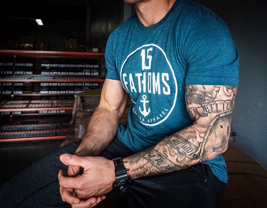Muscular male model wearing 15 Fathoms tee sitting down with hands clasped