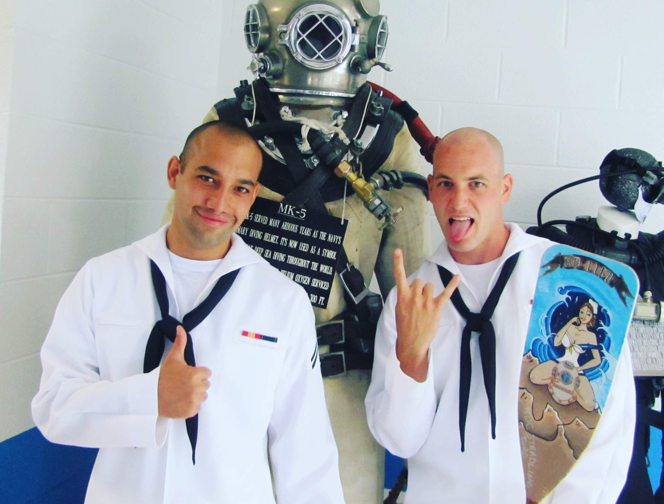 Two men in white navy service uniforms with a vintage diving suit and helmet in the background