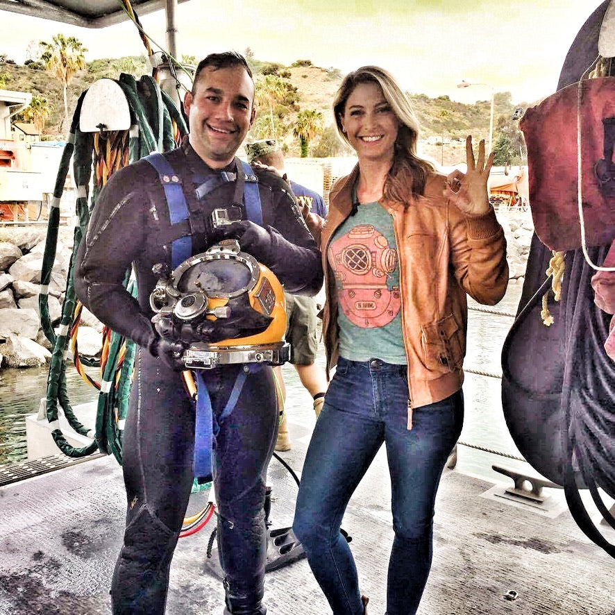 Two people, one in diving gear and the other in 15 Fathoms clothing, standing on a dock with diving equipment