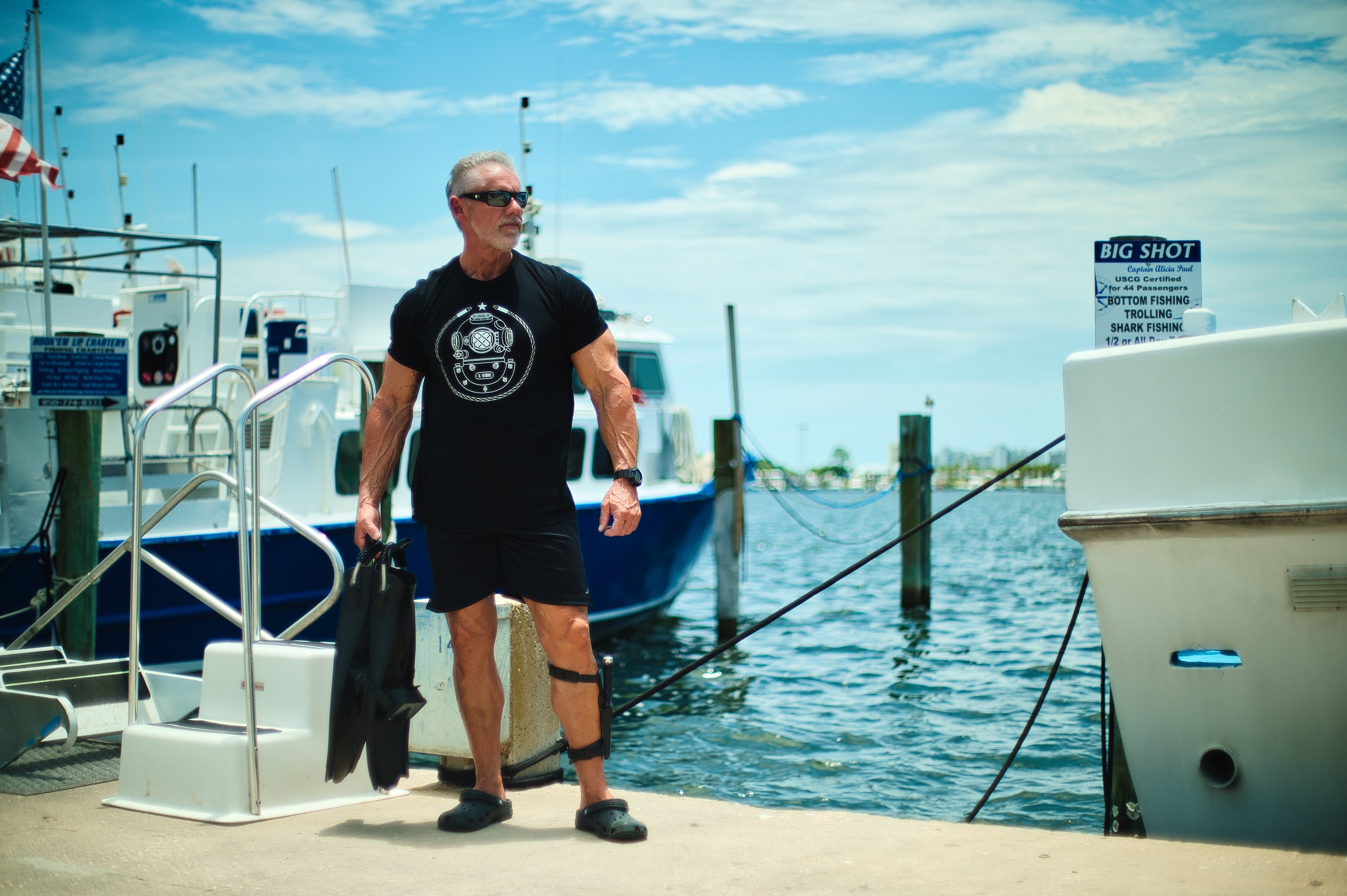 Man standing on a dock by boats wearing a black 15 Fathoms tee