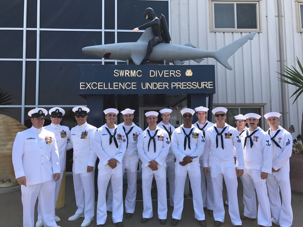 Group of men in navy sailor uniforms posing in front of a building with a shark sculpture and 'SWRMC Divers Excellence Under Pressure' sign.