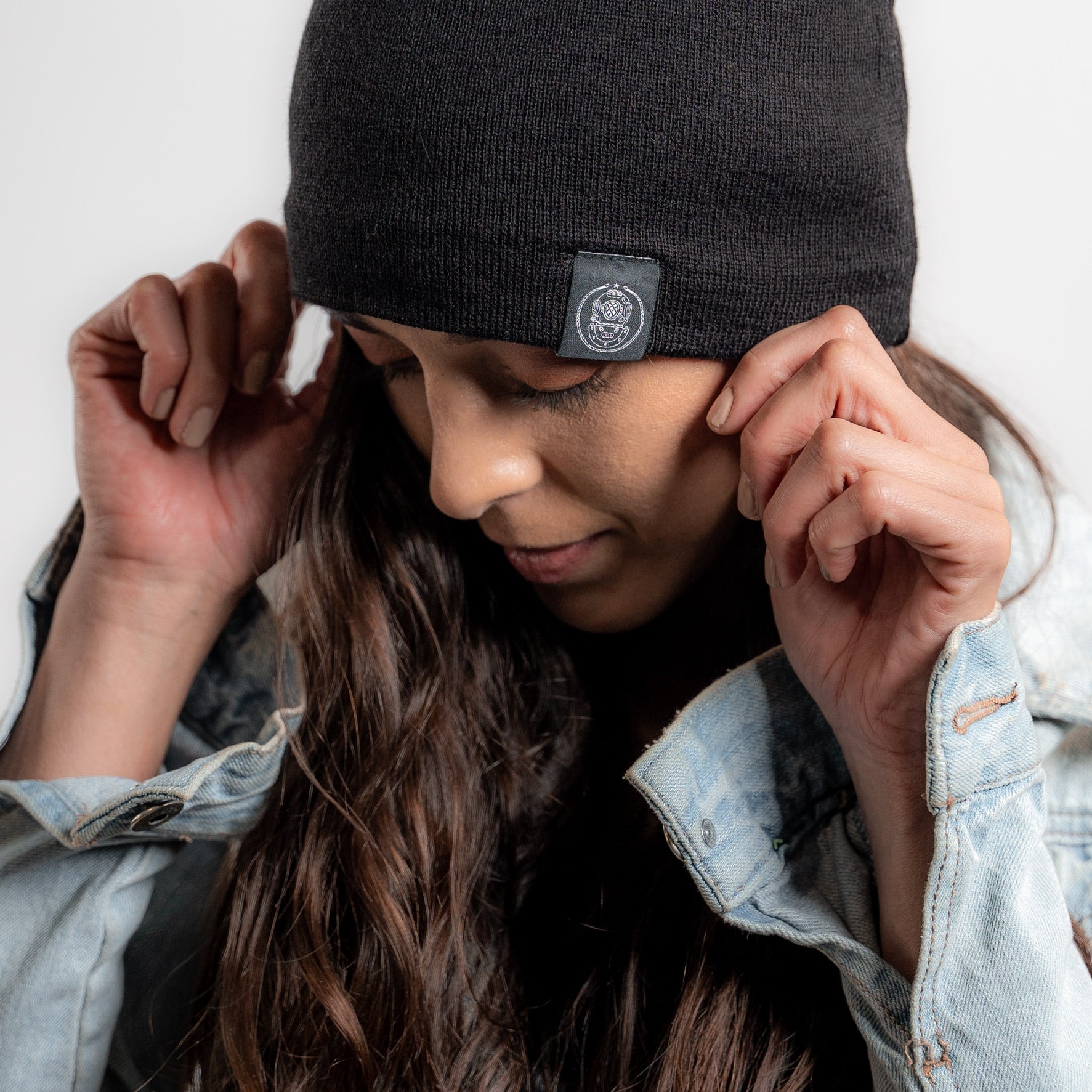 Woman wearing a black beanie with a 15 fathoms logo, adjusting it with both hands against a white background