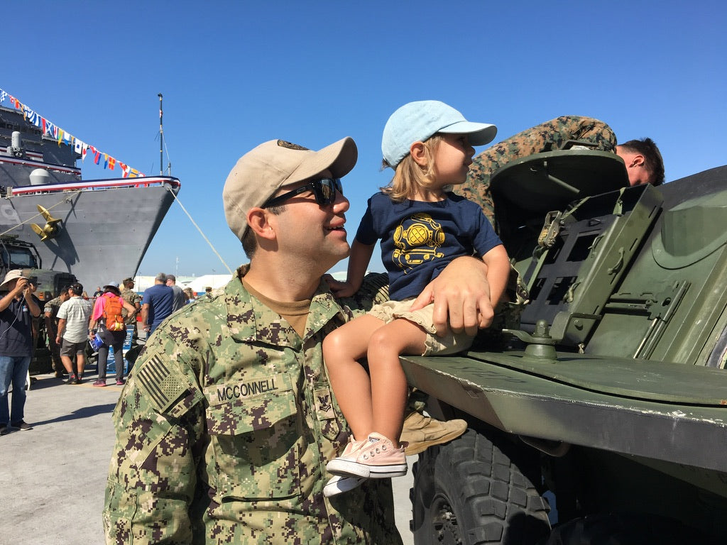 Man in military uniform holding a small child that's sitting on the side of a military vehicle