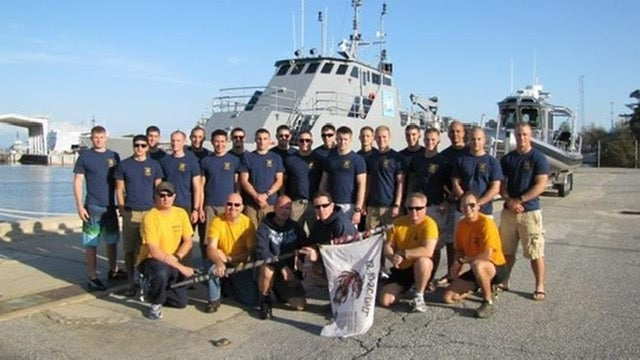 Group of men in navy uniforms posing on a dock with boats in the background