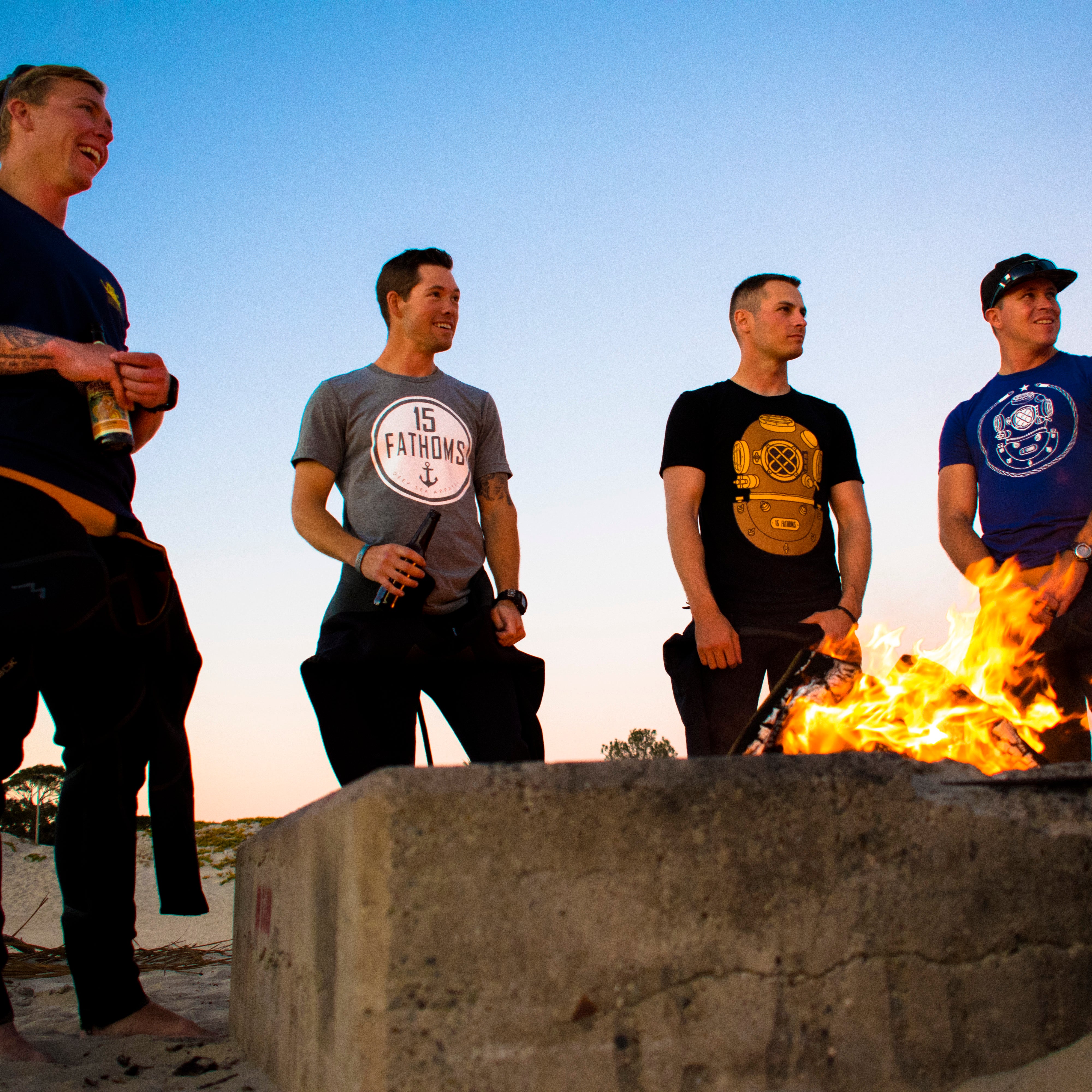 Four men gathered around a bonfire on the beach wearing 15 Fathoms tees.