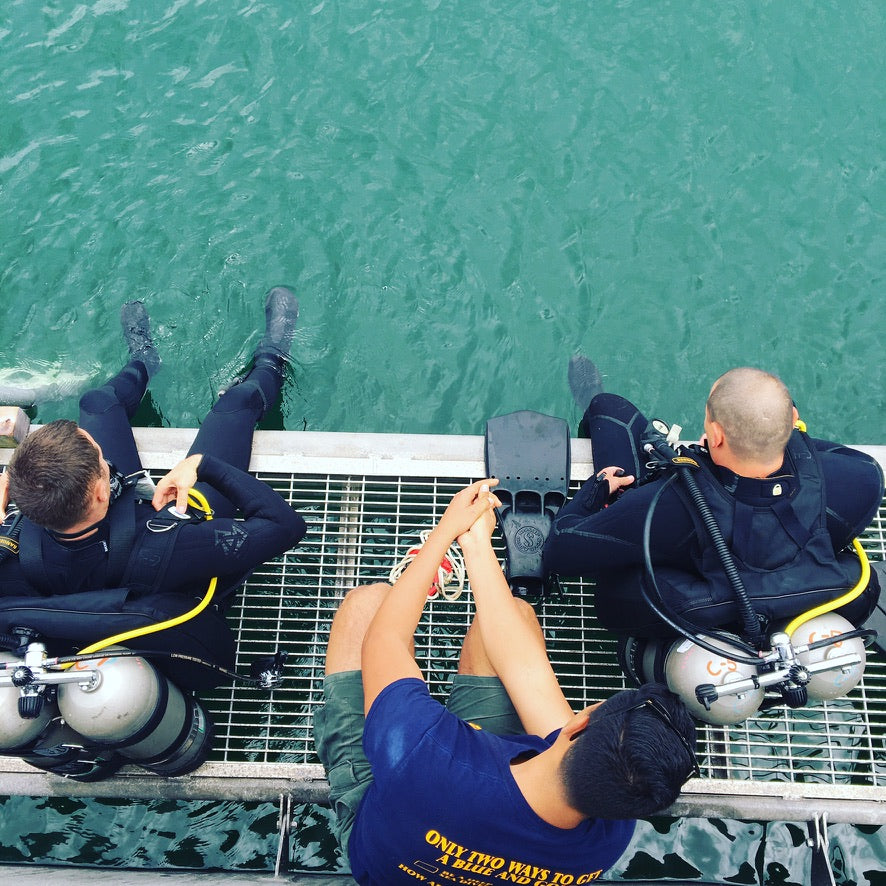 Two divers and one assistant sitting on a dock with feet in the water.