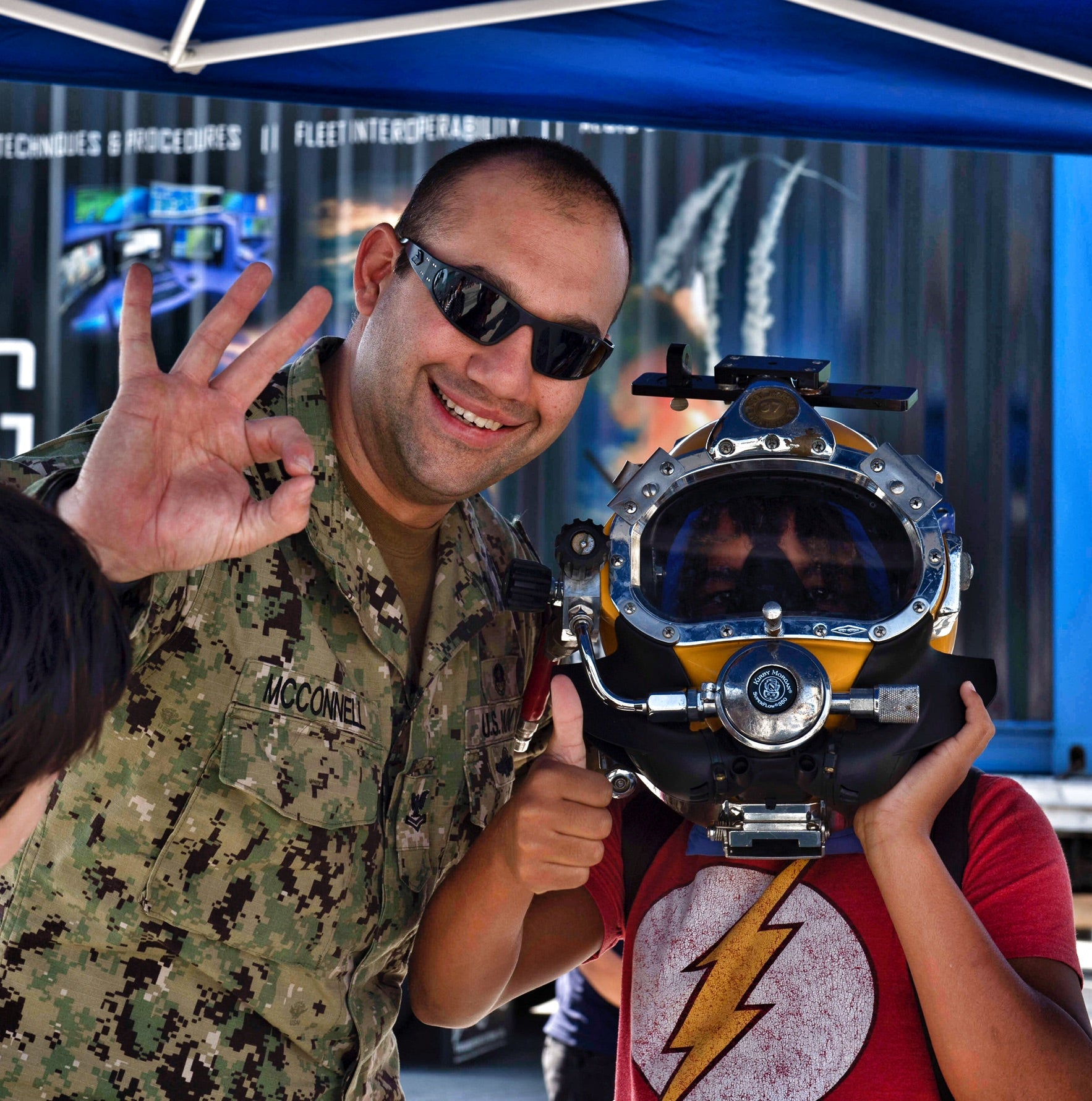 Man in military uniform with a kid wearing a diving helmet giving the O K signal and a thumbs up