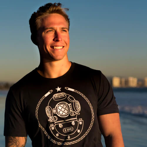 Man on the beach smiling into the sun wearing a black tee with white 15 fathoms diving helmet logo