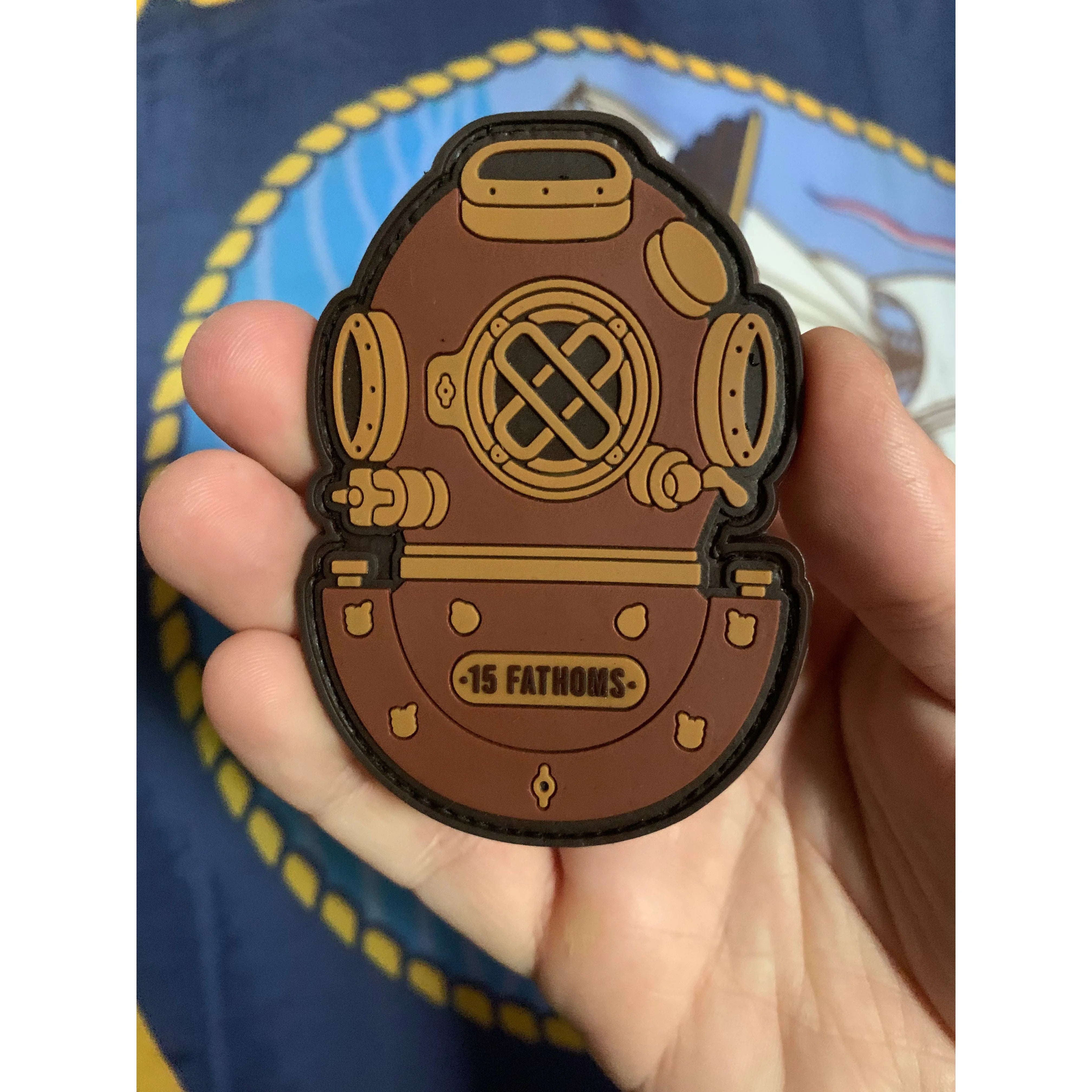 Mark-5 Patch - dive helmet with “15 Fathoms” text on base of helmet, someone holding up the patch